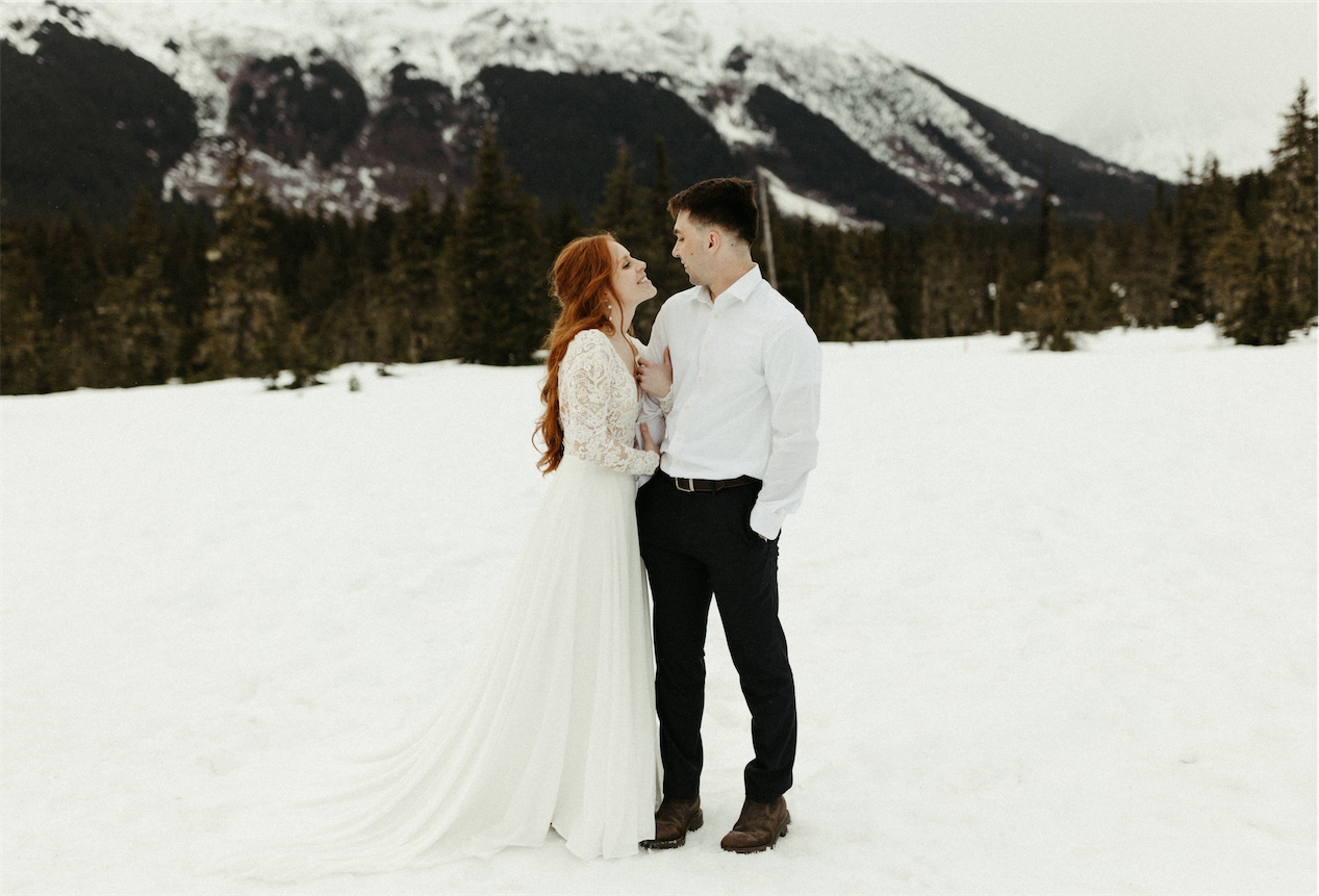 Couple holding one another while standing on a mountainside field covered in snow in Girdwood, Alaska