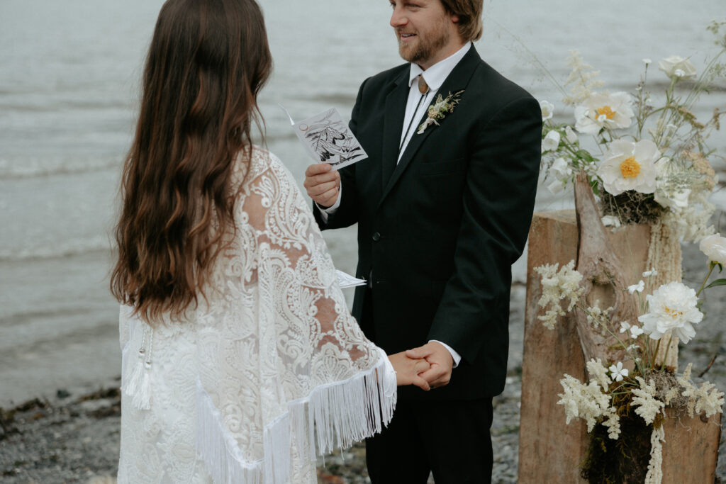 A man wearing a black suit holding a woman's hand while standing near the ocean.