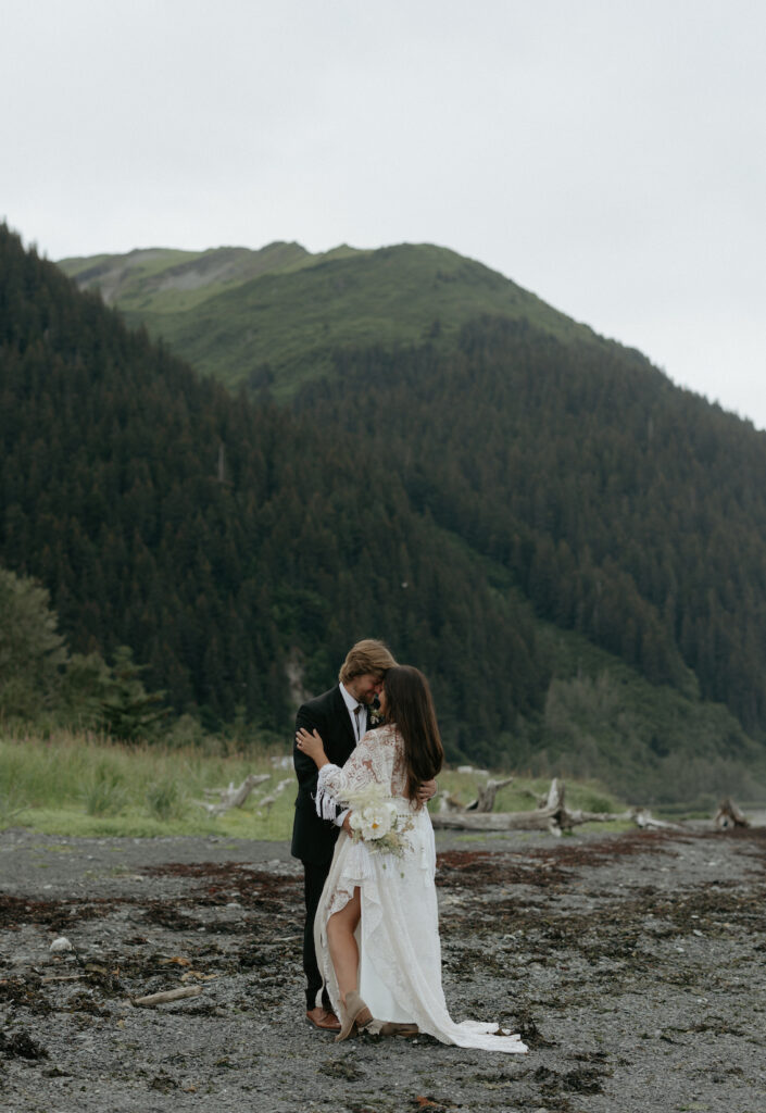 A couple embracing while standing on a beach near the mountains.