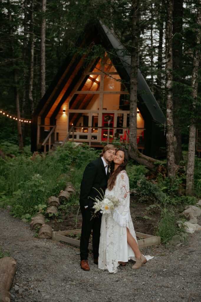 A couple standing with their cheeks smooshed together outside a lit up a-frame cabin. 