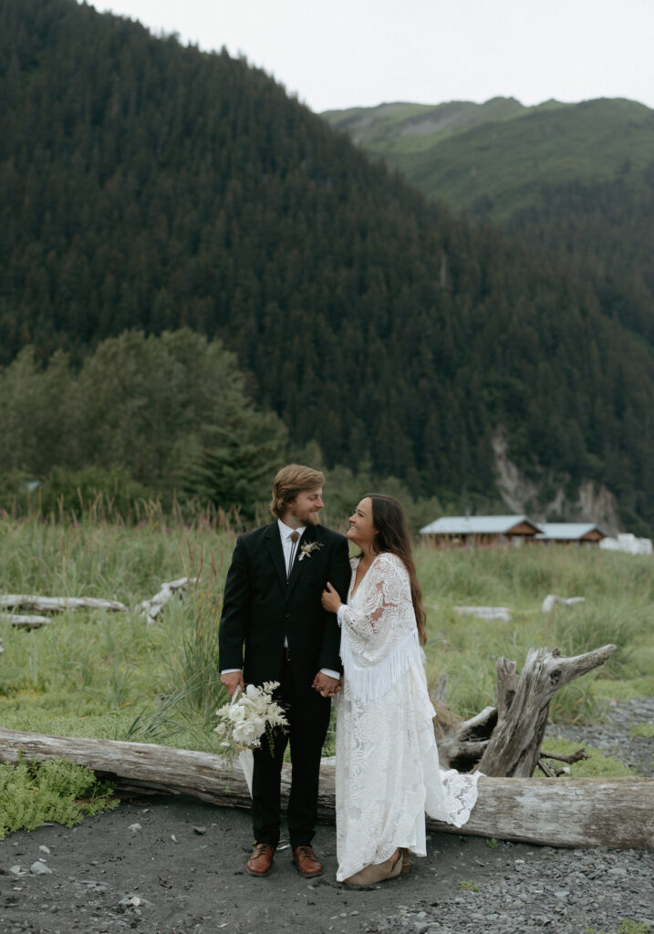 A couple looking at one another while standing next to a piece of driftwood. 