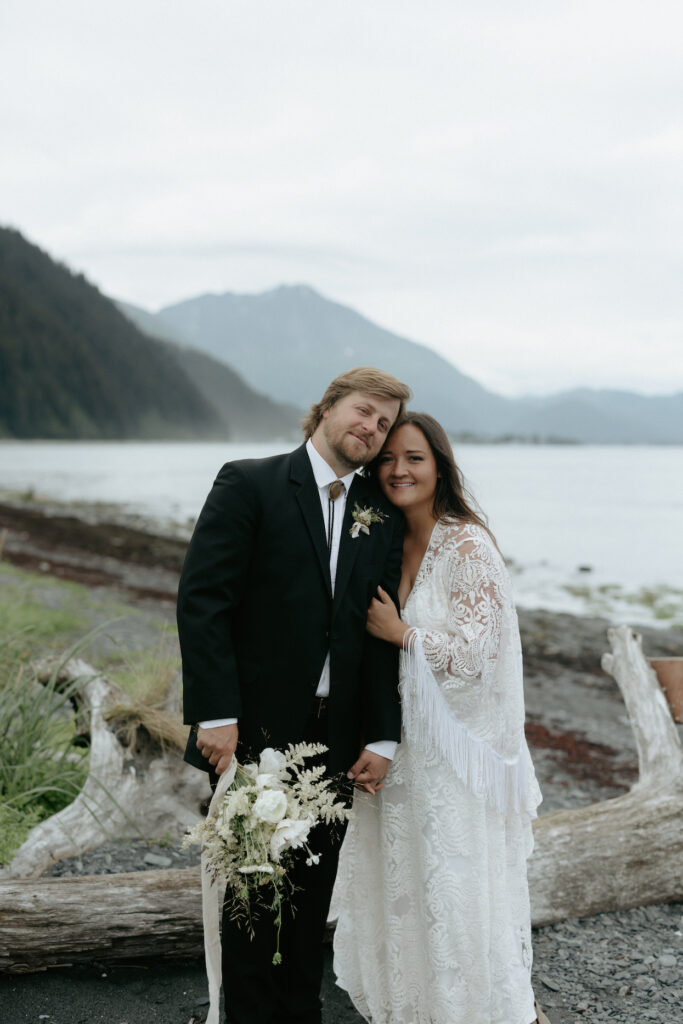 A couple snuggled up to one another while standing to a piece of driftwood. 