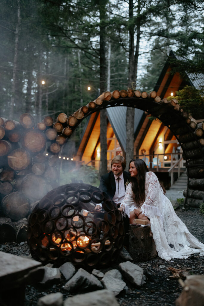 A couple outside their a-frame cabin sitting by the fire.