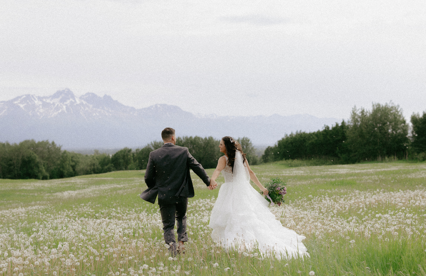 A couple holding hands and running through a field of dandelions during their spring wedding in Alaska.