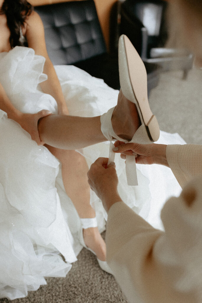 Mother tying her daughters shoes while she sits on a black couch wearing a wedding dress