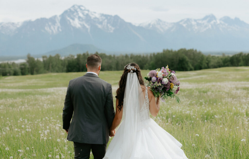 Couple walking in a field and heading toward the mountains while wearing wedding attire.