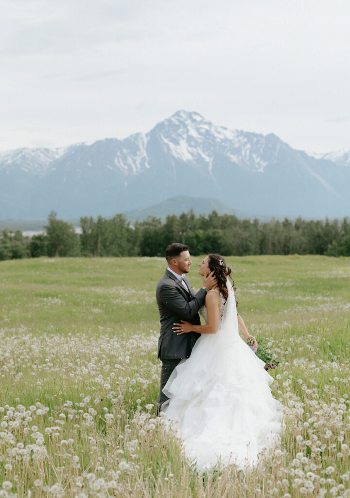 Couple wearing wedding attire holding onto one another in a field overlooking the mountains in Alaska