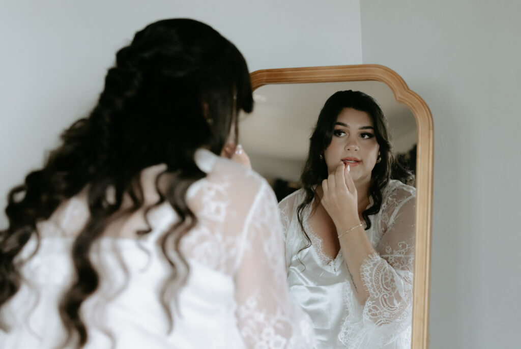 A woman wearing a white robe looking in a mirror and putting on lipstick.