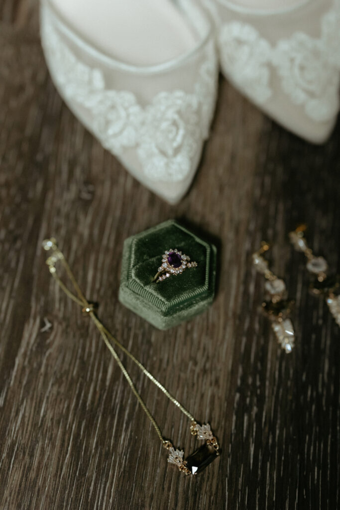 A green ring box with a purple stoned engagement ring in it. Surrounded by a pair of white shoes, a necklace and matching pair of earrings. 