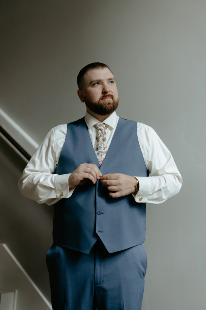 Man looking out the window as he buttons a blue vest. 