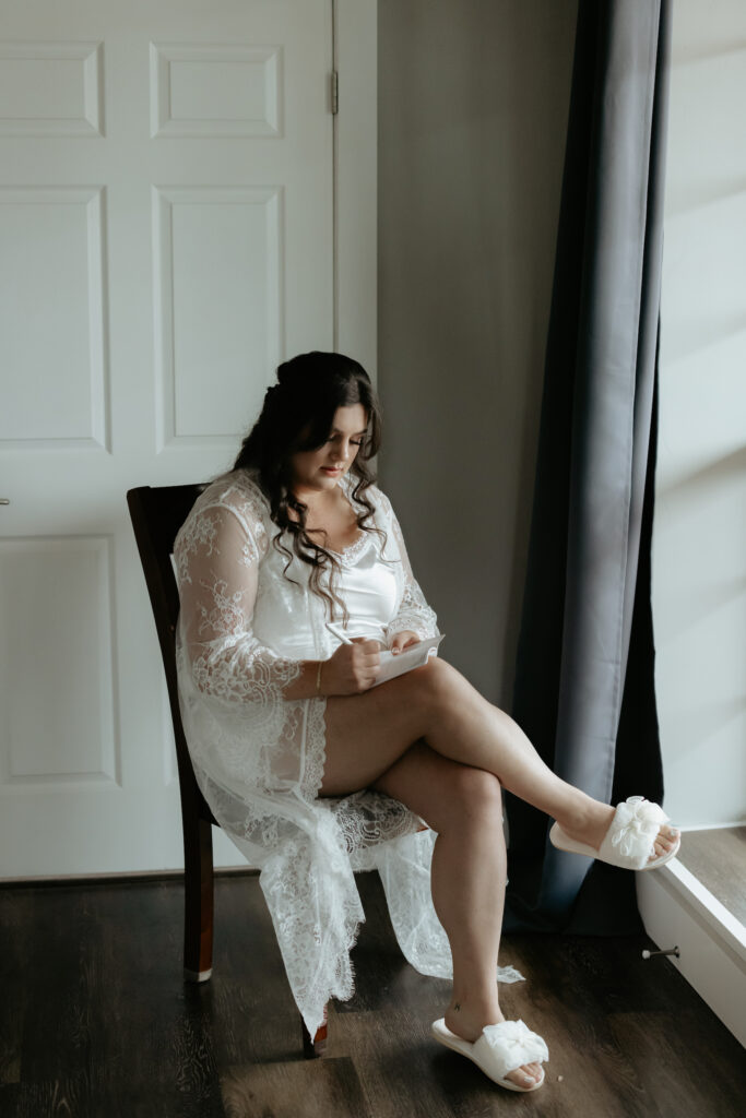 Woman sitting next to a window with blue curtains while wearing a bridal kimono and slippers.