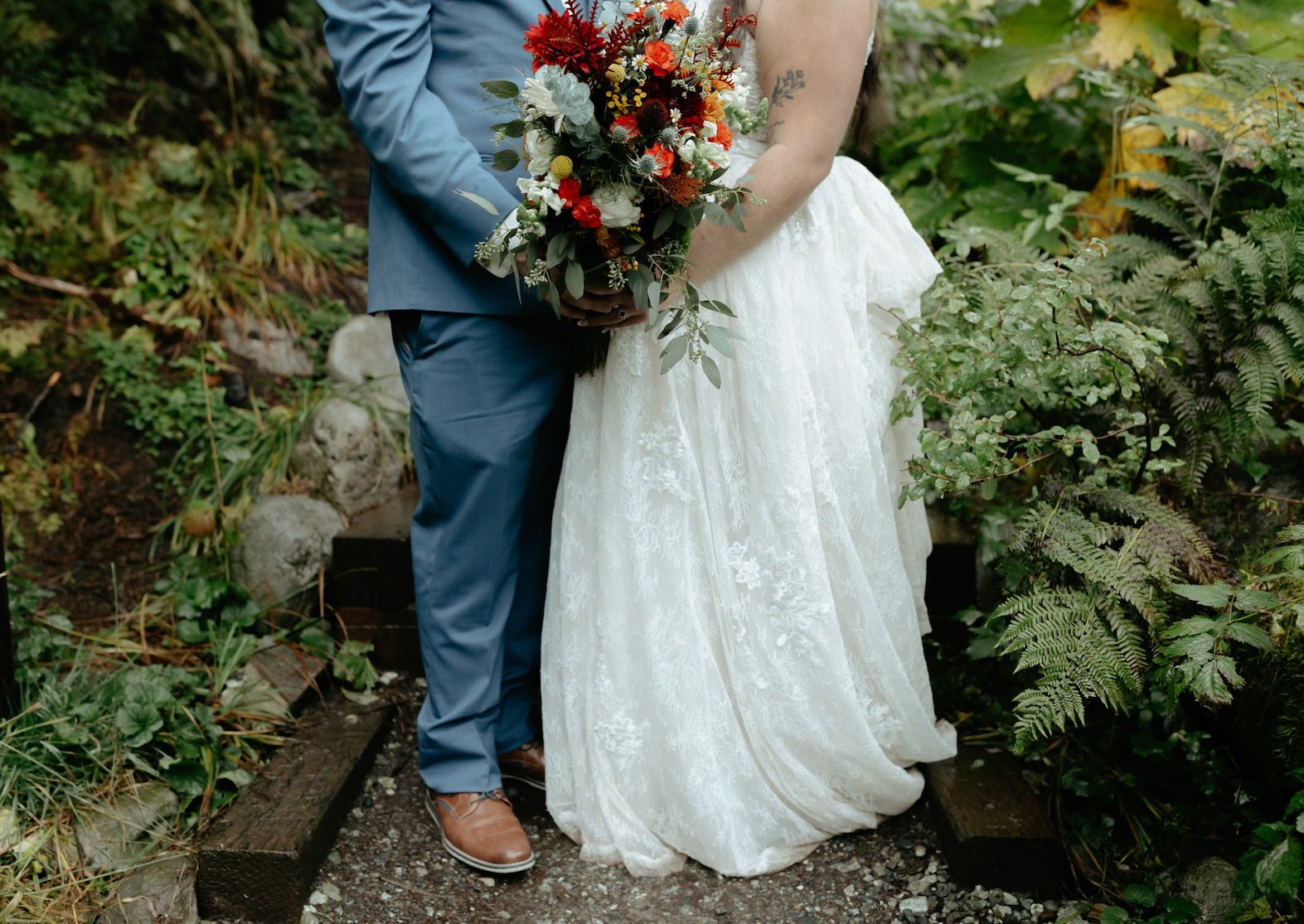 A couple holding a colorful bouquet of florals while surrounded by lush fall foliage near a waterfall in Alaska. 