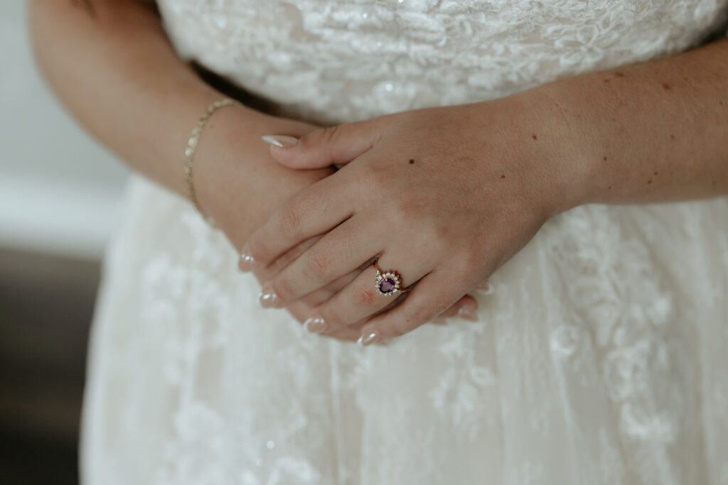 Woman standing with hands in front of her while wearing a purple stoned engagement ring.