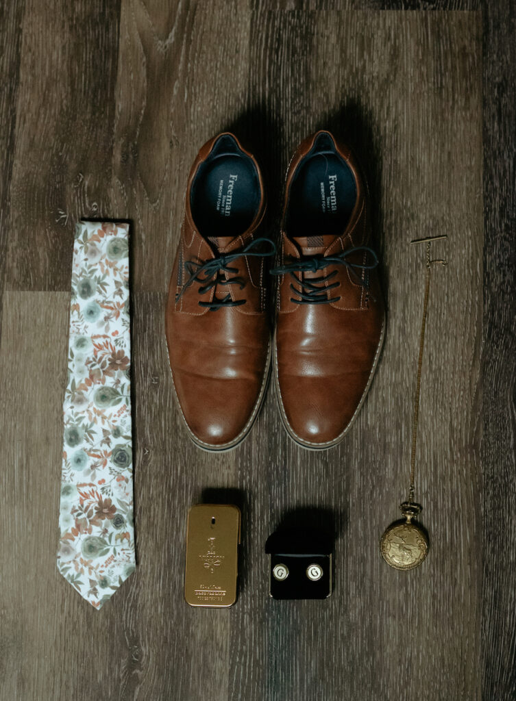 Mans shoes, cologne, cuff links and a shoes sitting on a dark floor.