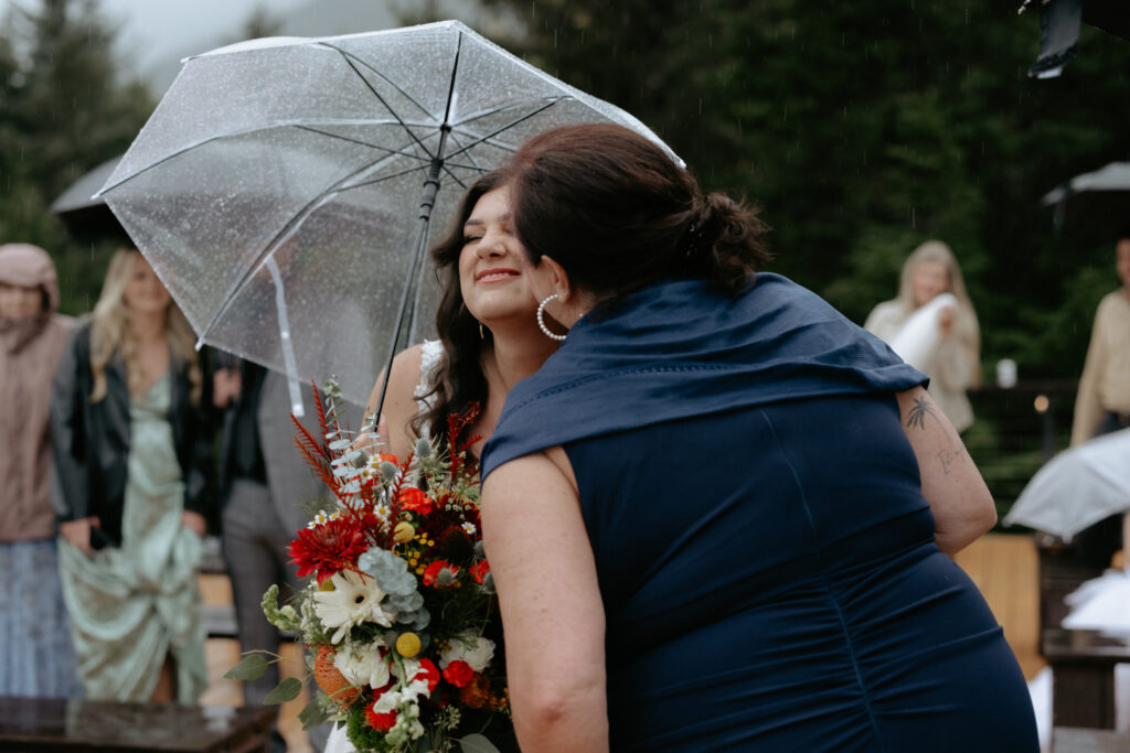 Mother kissing her daughter on the cheek as she walks down the aisle holding a colorful bouquet. 