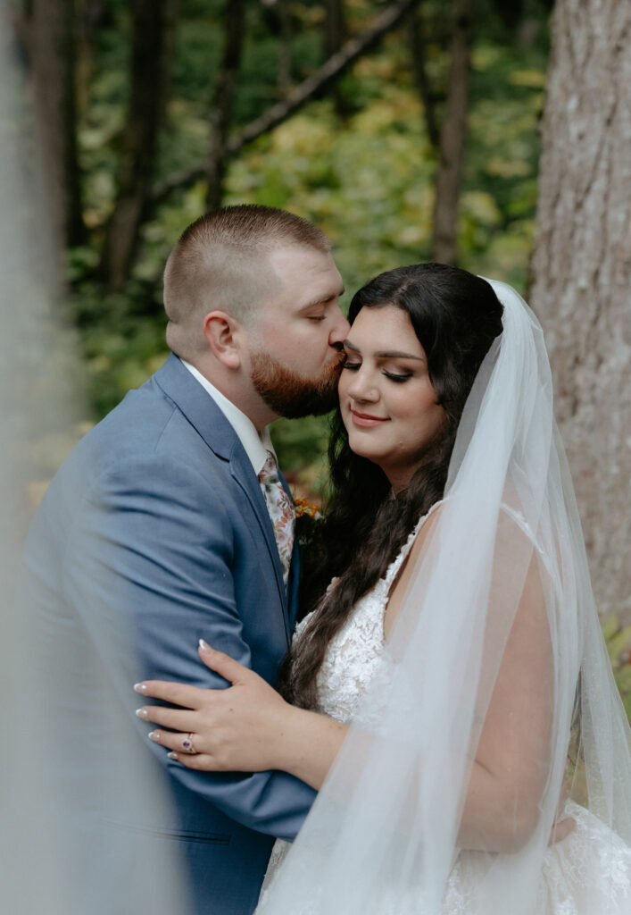 Man kissing woman's forehead while standing in a lush forrest 