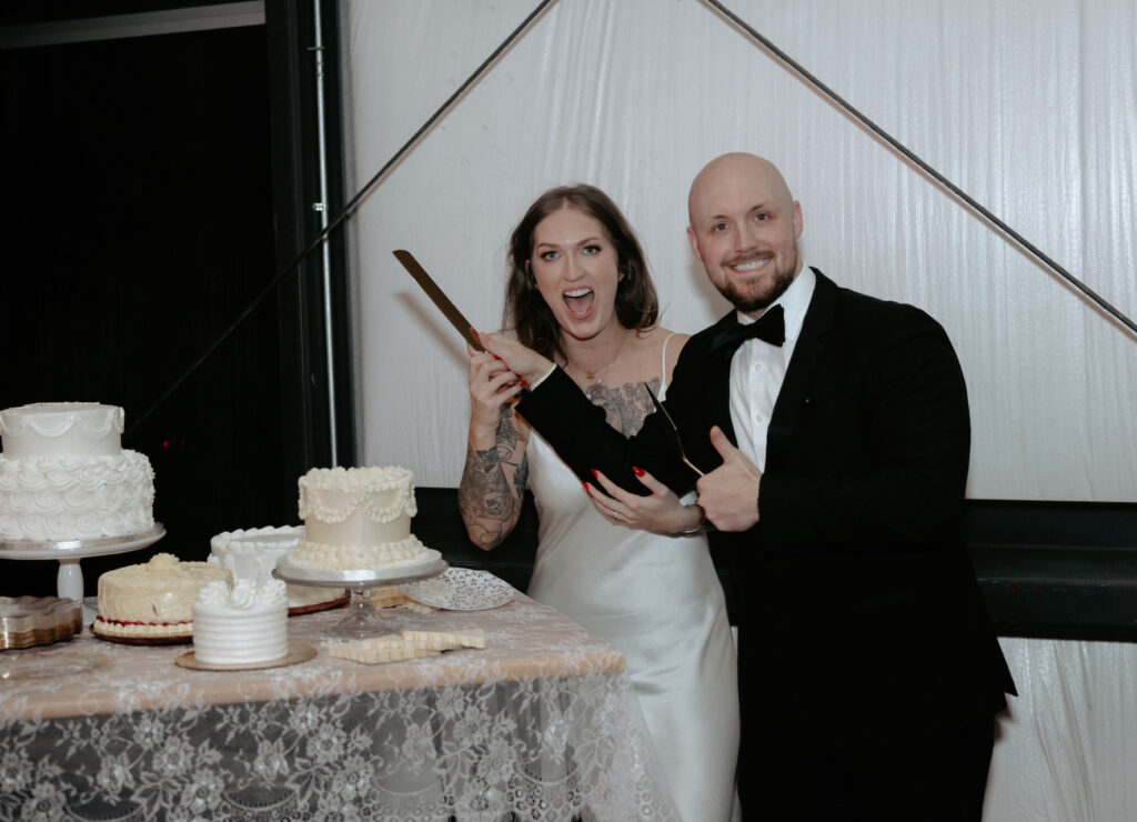 A couple holding a knife and standing next to their wedding cake. 