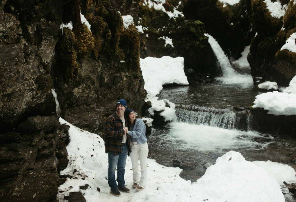 Couple holding hands while stand next to a snow covered waterfall.