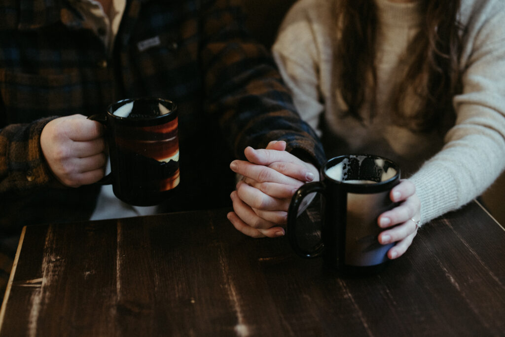 A couple holding hands while holding onto black coffee mugs. 