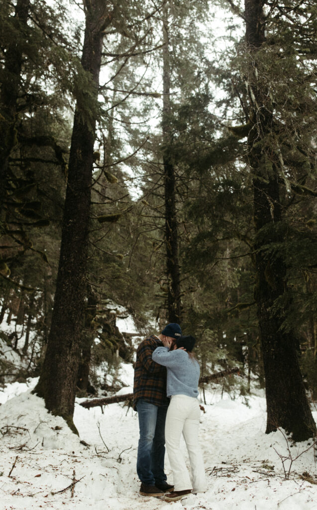 A couple embracing one another while standing in a snowy forrest.