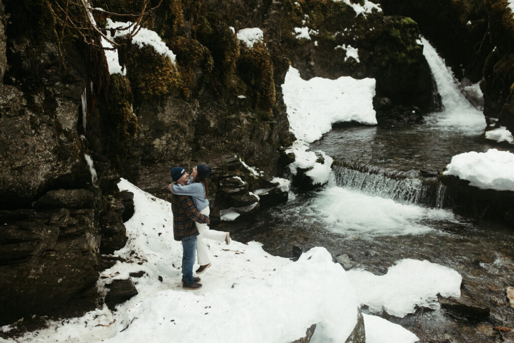 Man picking up woman while standing next to a snowy waterfall. 