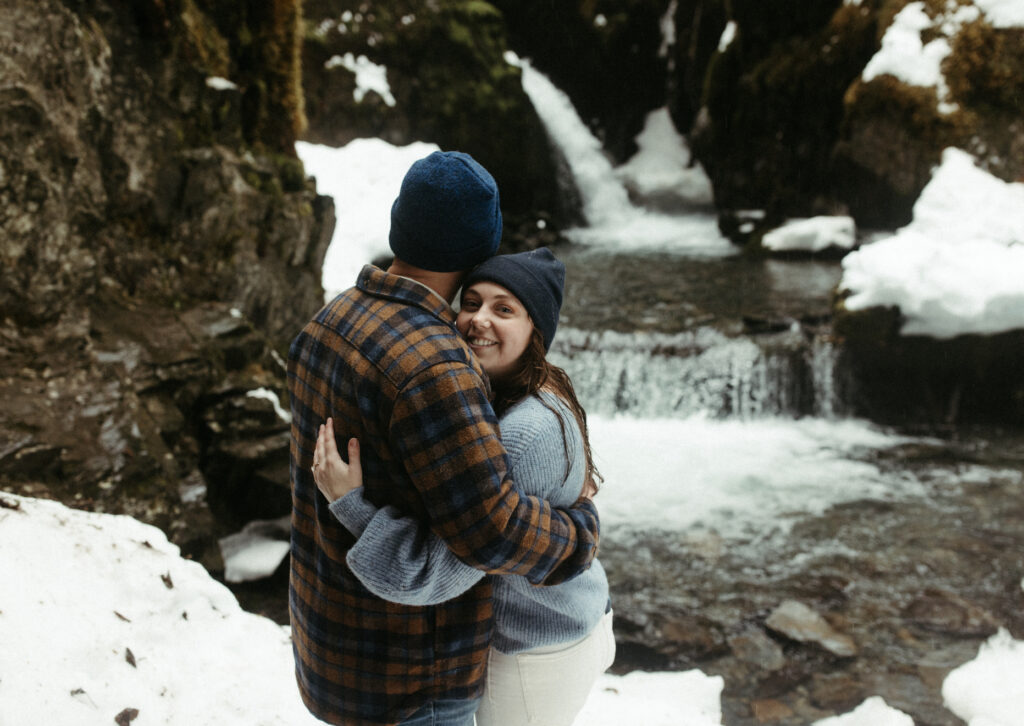 A couple snuggled together next to a waterfall. 