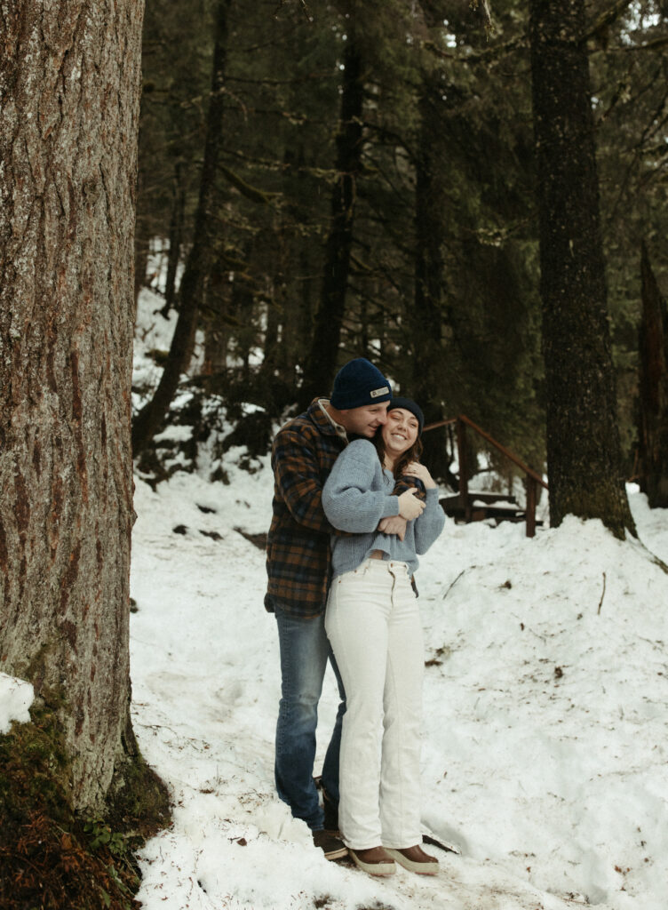 A man hugging a woman from behind while in a snowy forrest. 