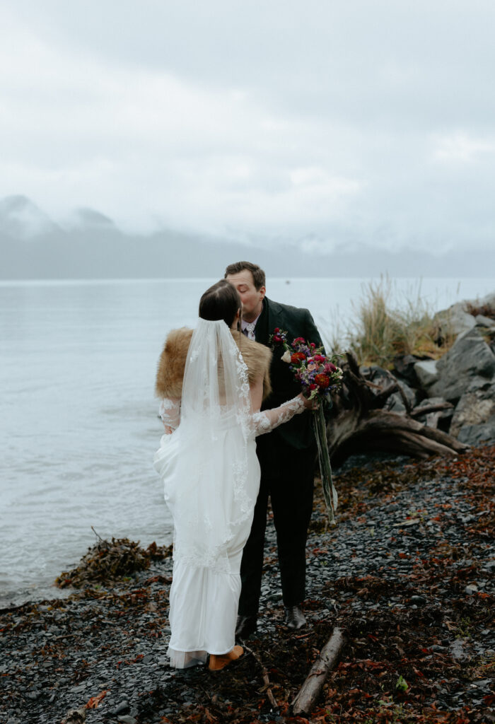 A couple kissing one another while standing on the beach next to the mountains.