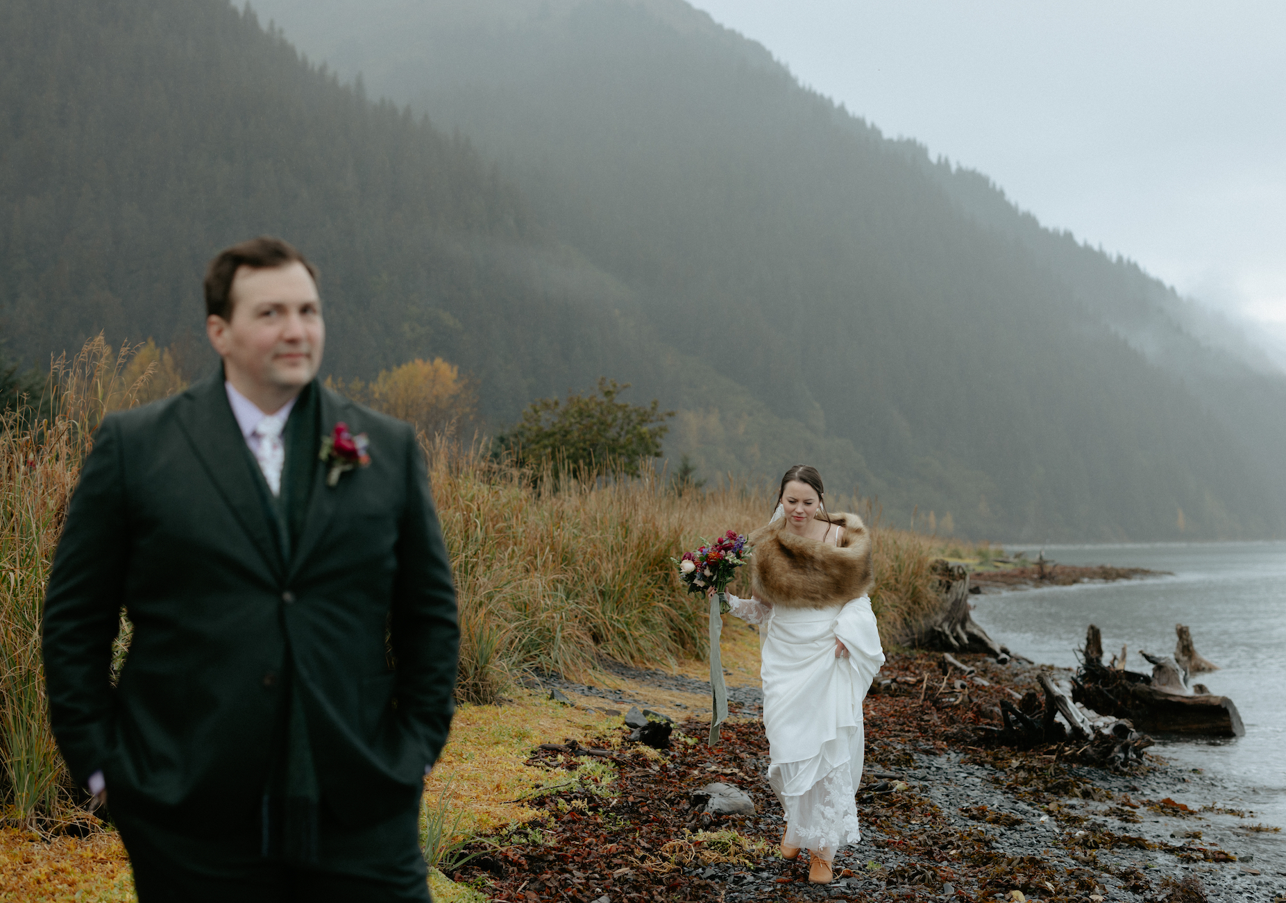 Woman wearing a white dress walking along a beach in Seward toward a man in a green suit. She is getting ready for a wedding first look at Salted Roots a wedding venue in Alaska. 