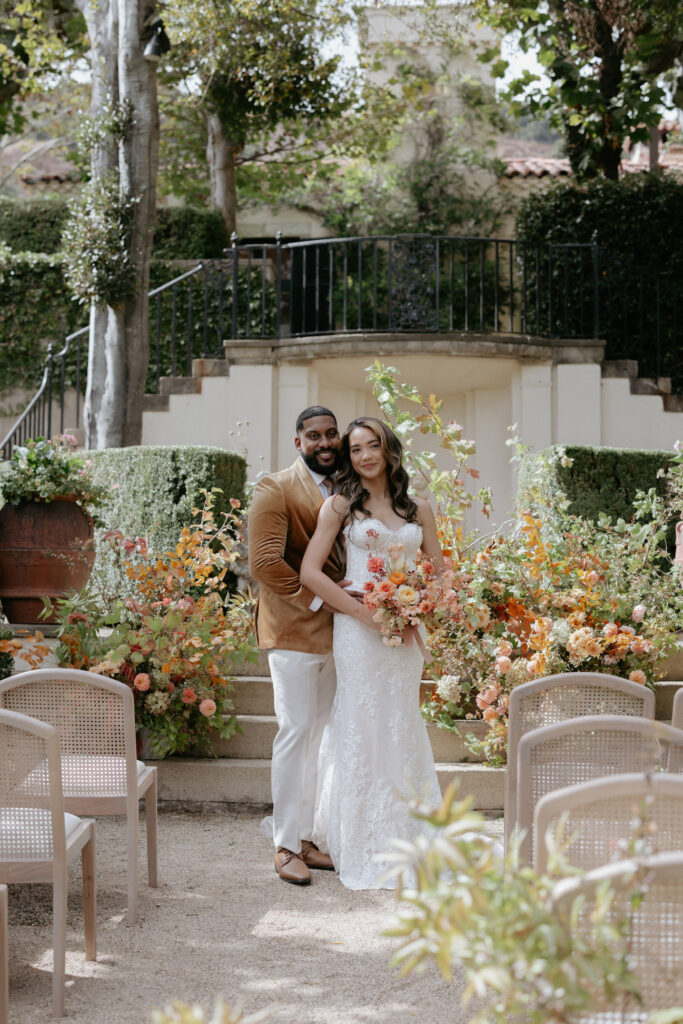 A couple standing in front of a large floral arch while surrounded by white seated chairs. 