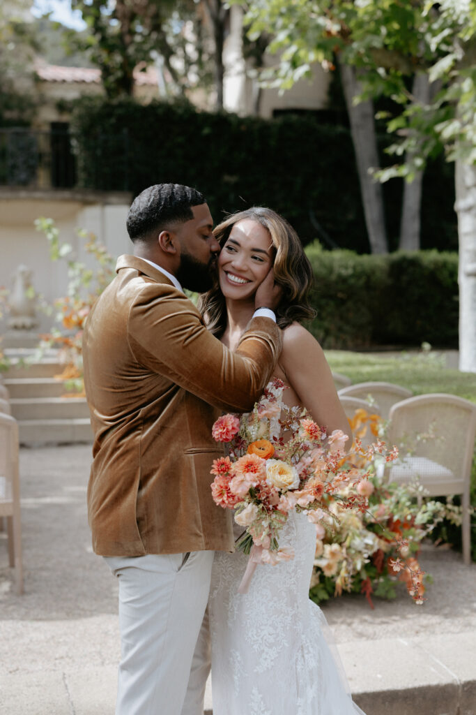 A man kissing a woman's forehead as she is holding onto her flowers.