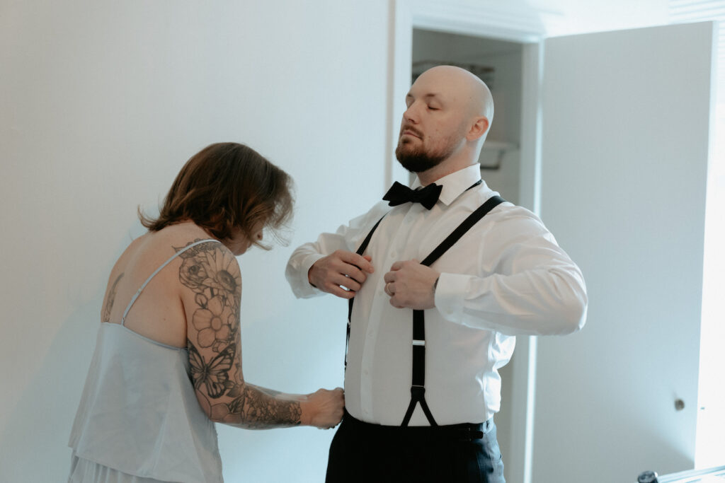 A woman helping a man get dressed by adjusting his suspenders. 