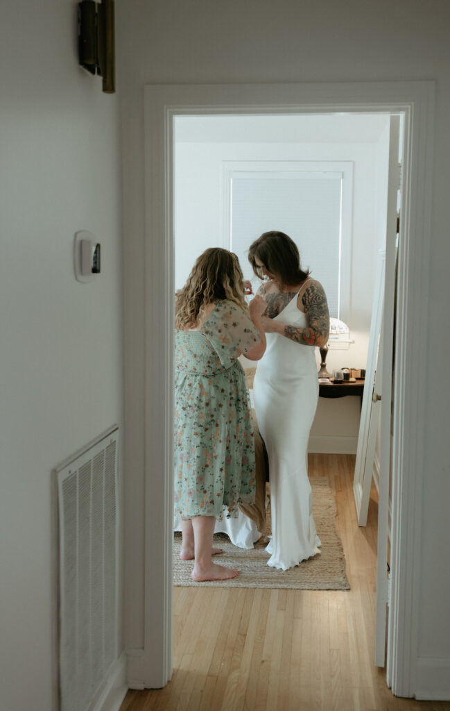 A woman wearing a floral dress helping a woman in a white dress put on a piece of jewlry. 