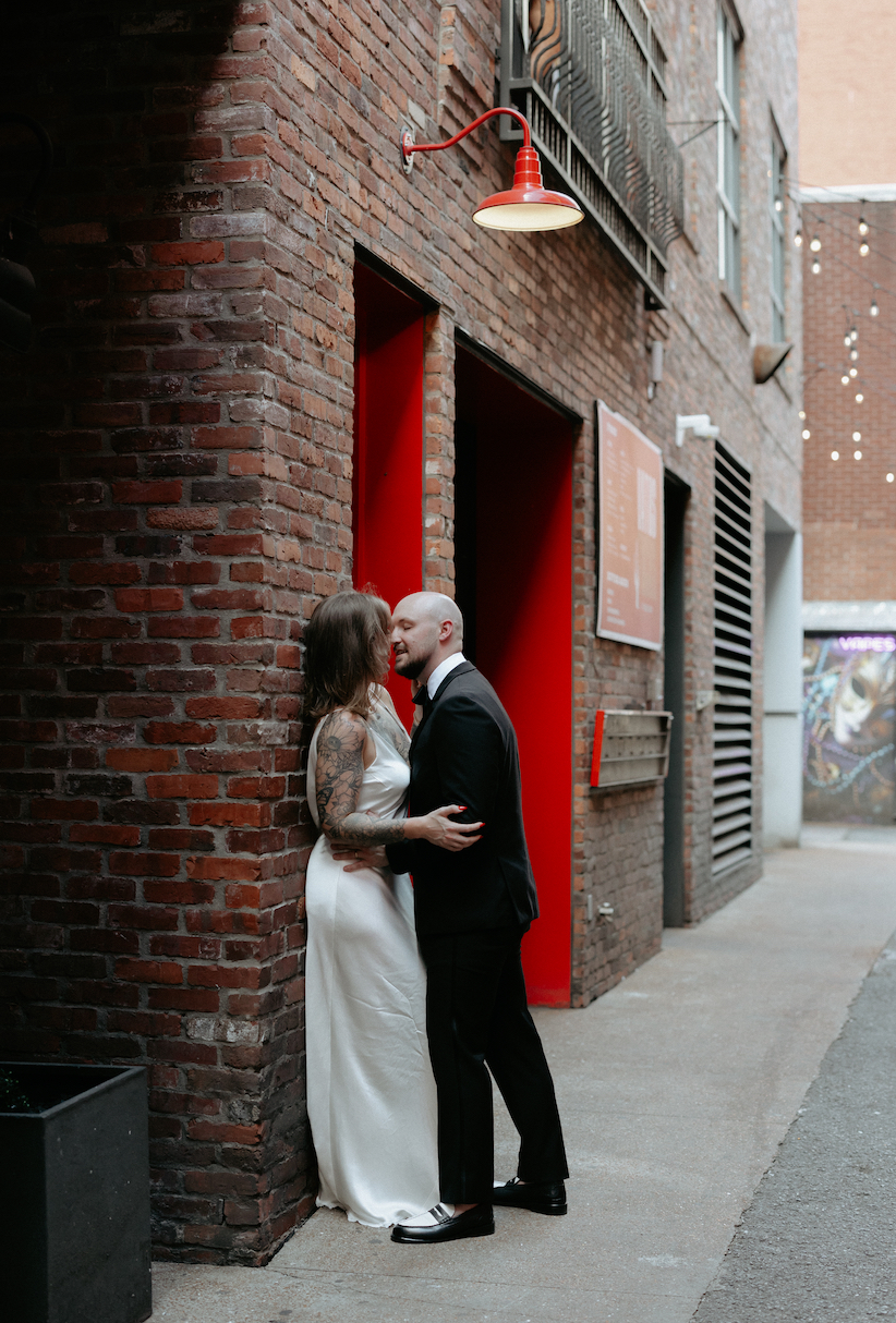 A couple embraced with one another while standing against a brick wall. 