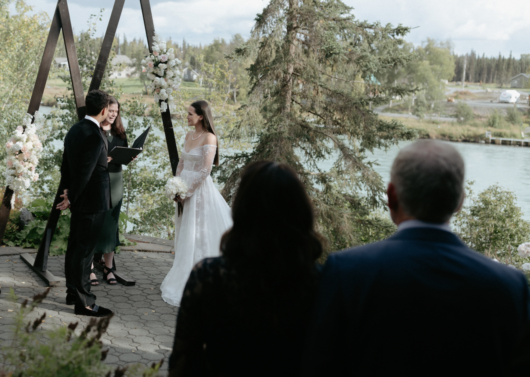 A couple standing next to a wooden arch while surrounded by family and friends.