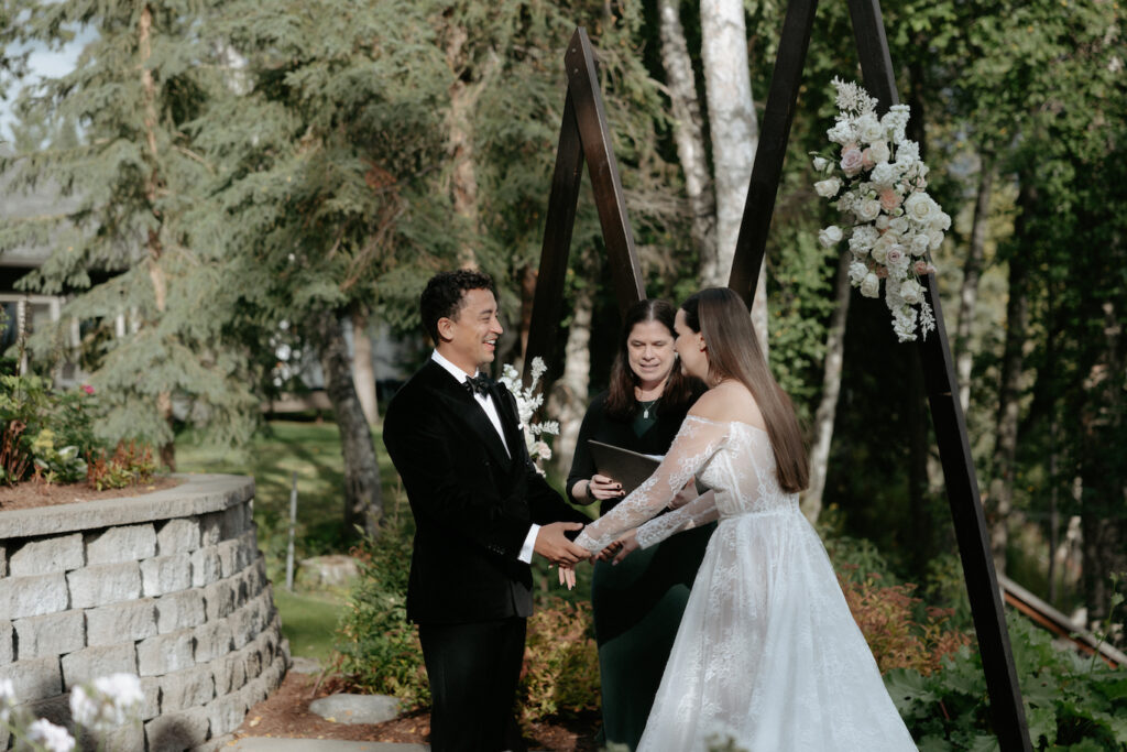 A couple holding hands in front of a wooden arch with pink and white florals attached to it. 