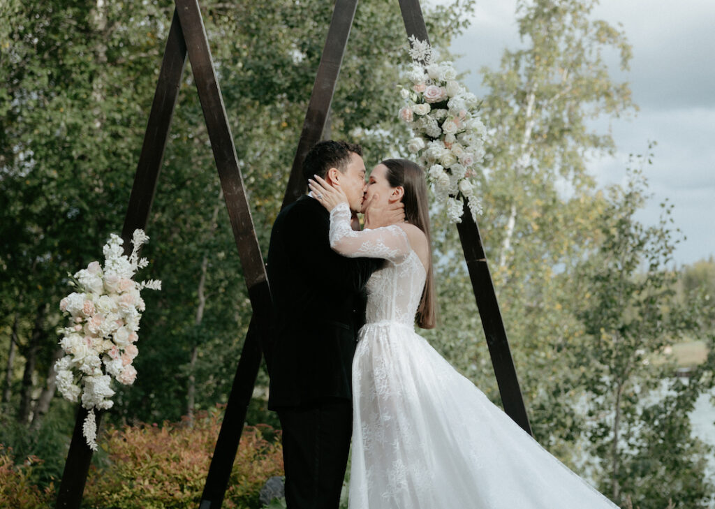 A couple kissing while standing in front of a wooden arch.