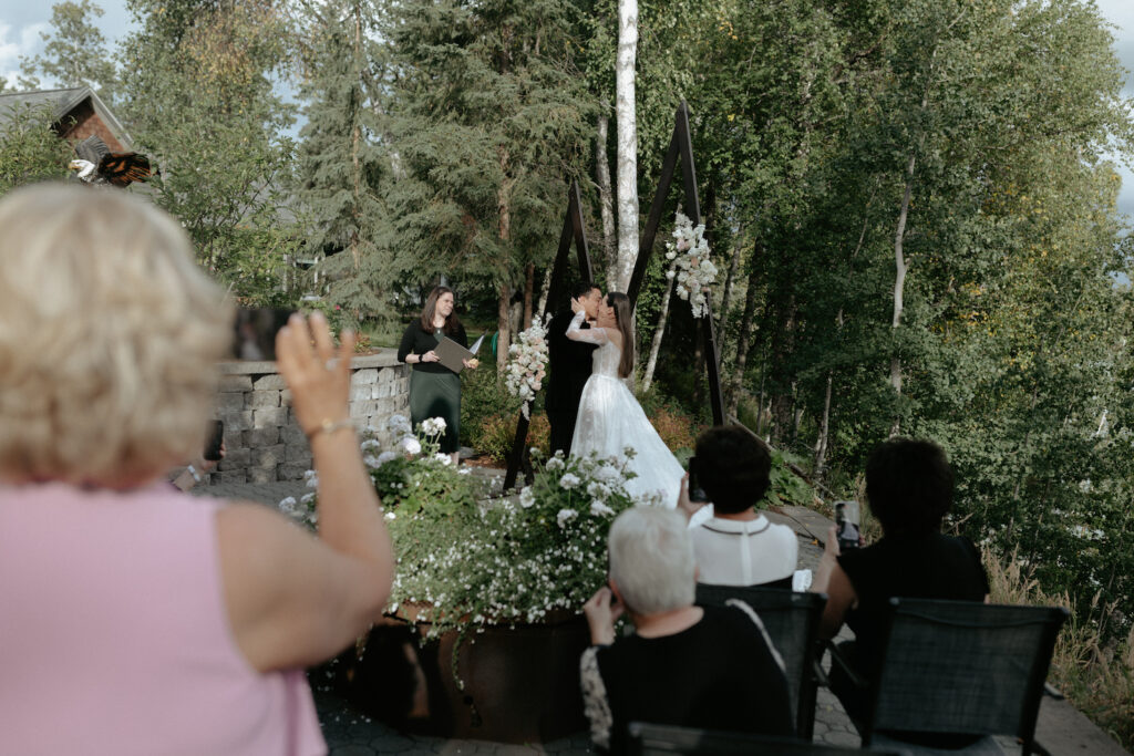 A couple wearing wedding attire kissing while surrounded by family and friends.