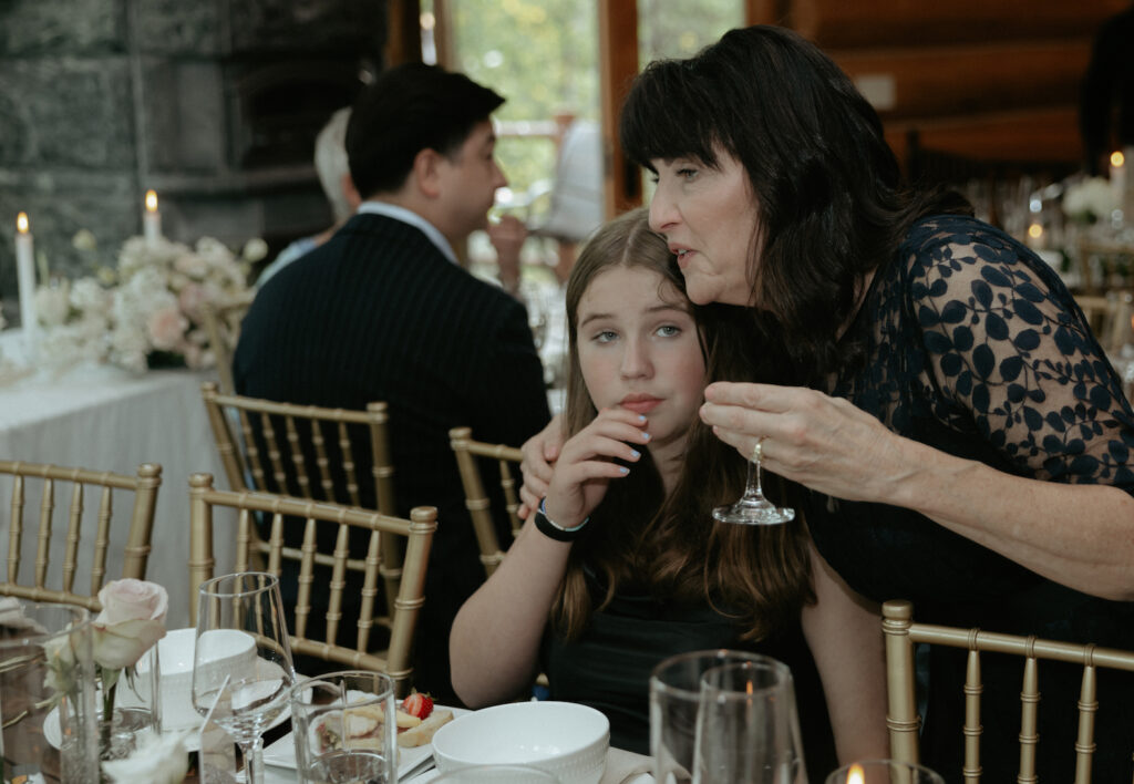 The mother of the bride holding a drink and hugging a girl that is sitting in a chair. 