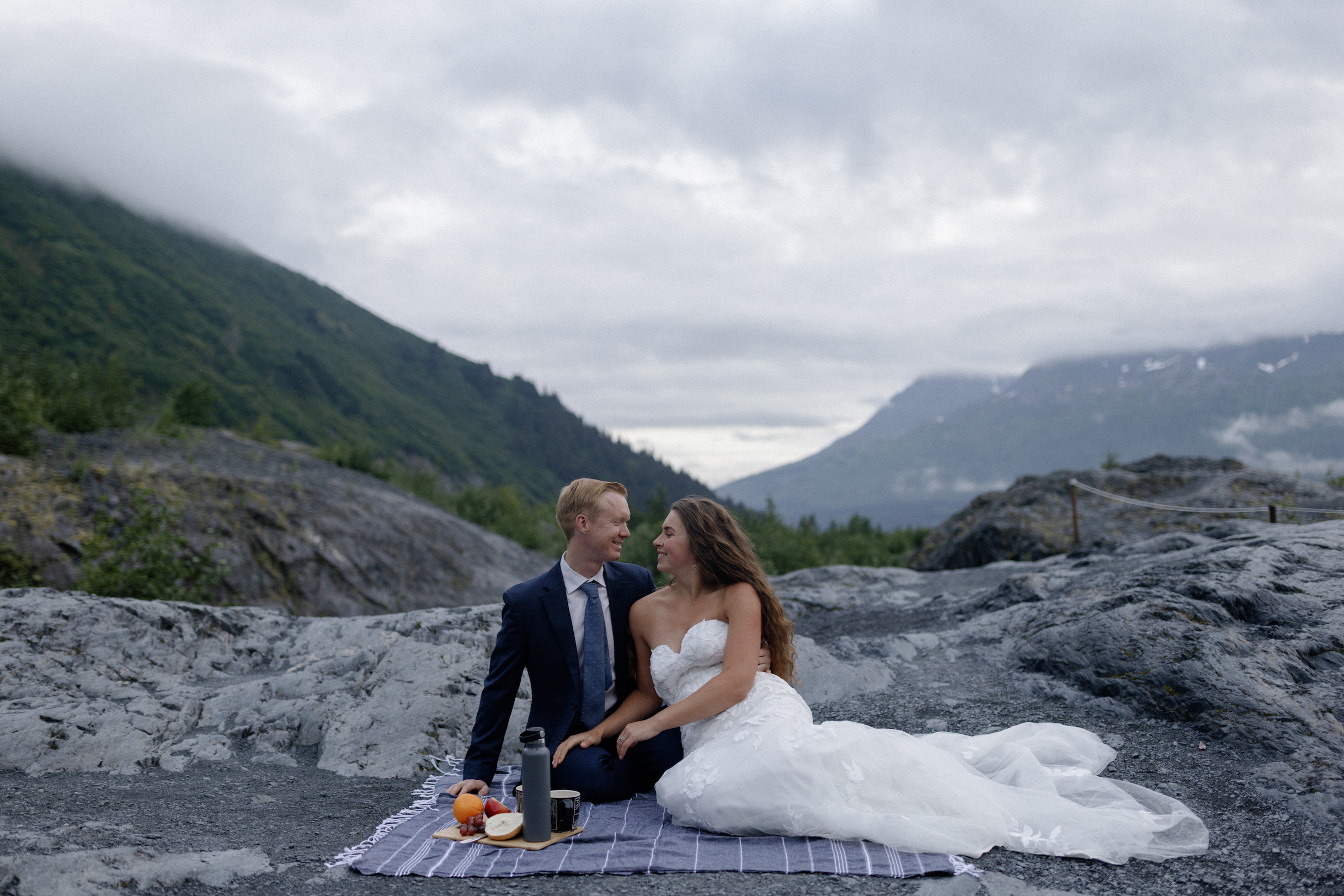 A bride and groom sitting on a blanket and eating. 