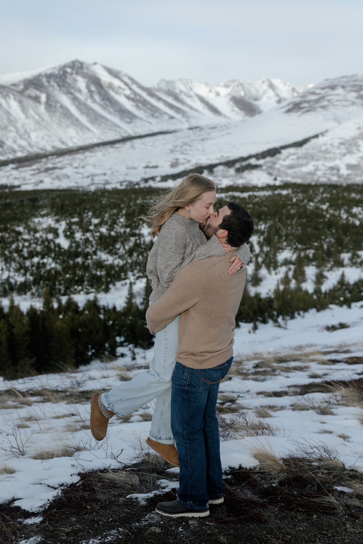 A man picking up a woman while standing in front of a snowy mountain.